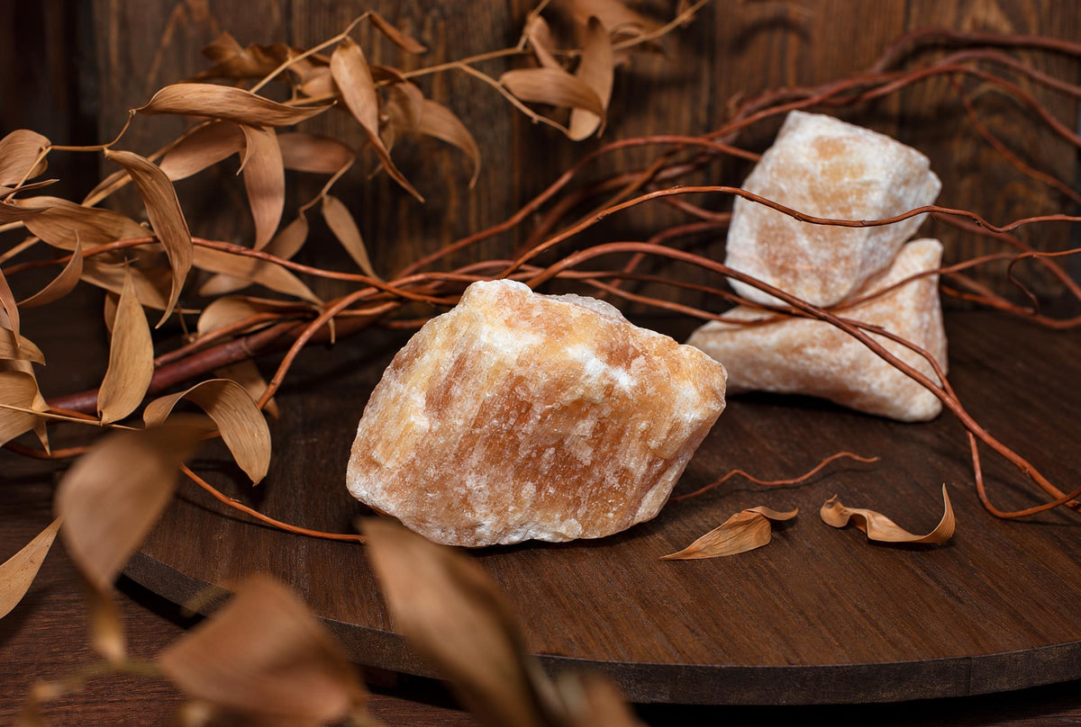 Three raw calcite crystals, orange and white, rest on a dark wooden surface with dried leaves and twigs.