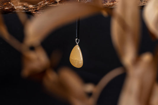 A teardrop-shaped, pale orange pendant hangs from a black cord against a dark background with blurred wooden elements.