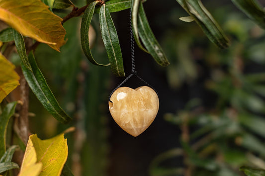 A polished, heart-shaped calcite pendant hangs from a black cord against green leaves and a dark background.