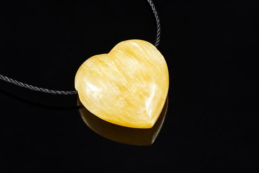 A polished heart-shaped Calcite pendant in pale yellow, resting on a dark surface with a black cord.