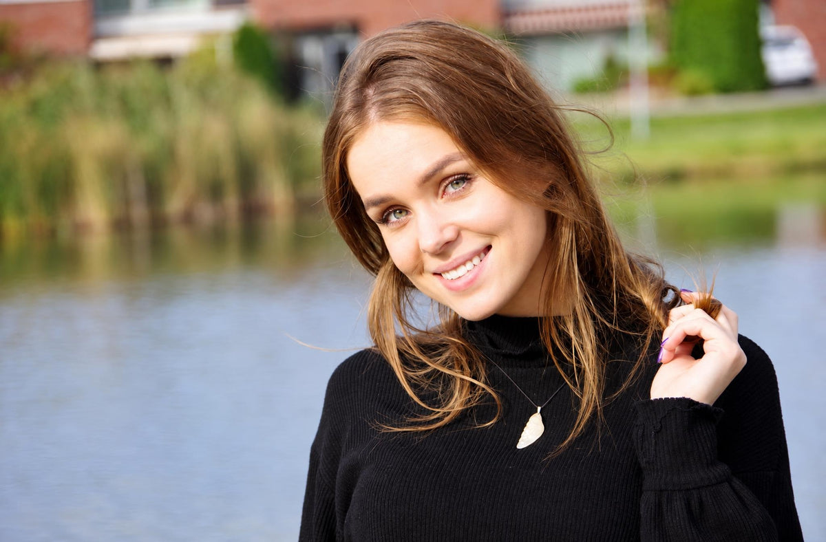 A smiling woman with brown hair wears a black turtleneck and a light-coloured wing-shaped pendant on a thin chain, holding her hair near a lake.