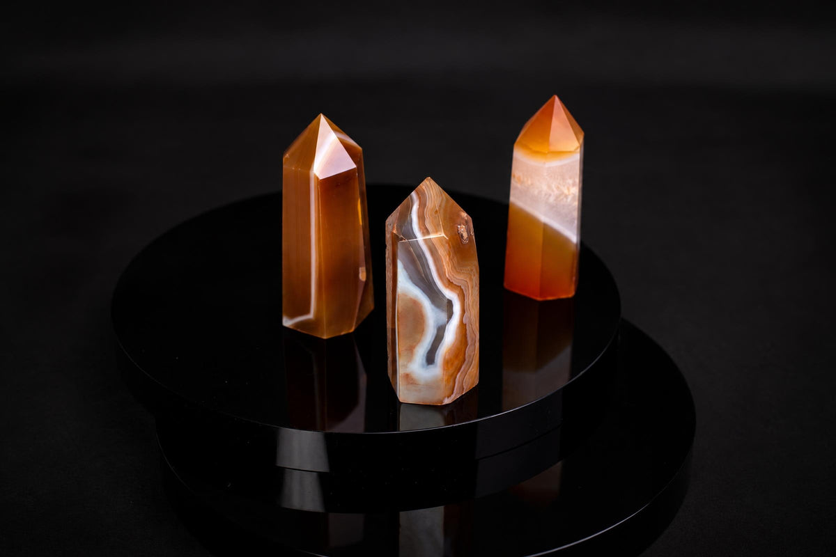 Three polished carnelian crystal points with orange and white banding stand on a dark, reflective surface.