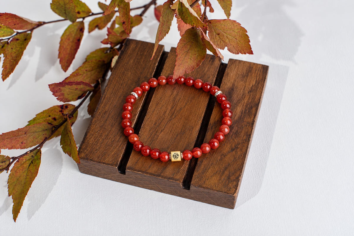 A red beaded bracelet with a gold-toned square charm rests on a dark wooden surface, surrounded by autumn leaves.