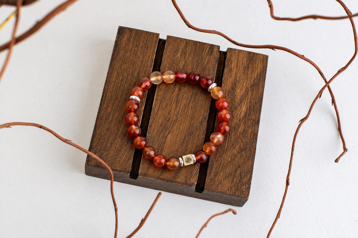 A beaded bracelet with red and orange spheres, silver-toned spacers, and a gold-coloured square charm rests on a dark wooden surface.