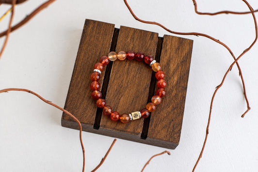 A beaded bracelet with red and orange spheres, silver-toned spacers, and a gold-coloured square charm rests on a dark wooden surface.