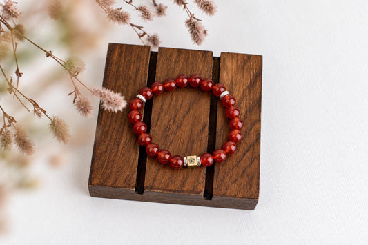 A red beaded bracelet with silver-toned accents rests on a dark wooden surface, surrounded by dried flowers.