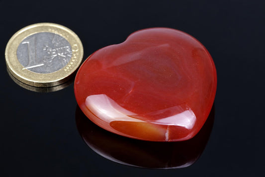 A polished red heart-shaped stone with white and orange bands sits on a dark surface next to a one-euro coin.