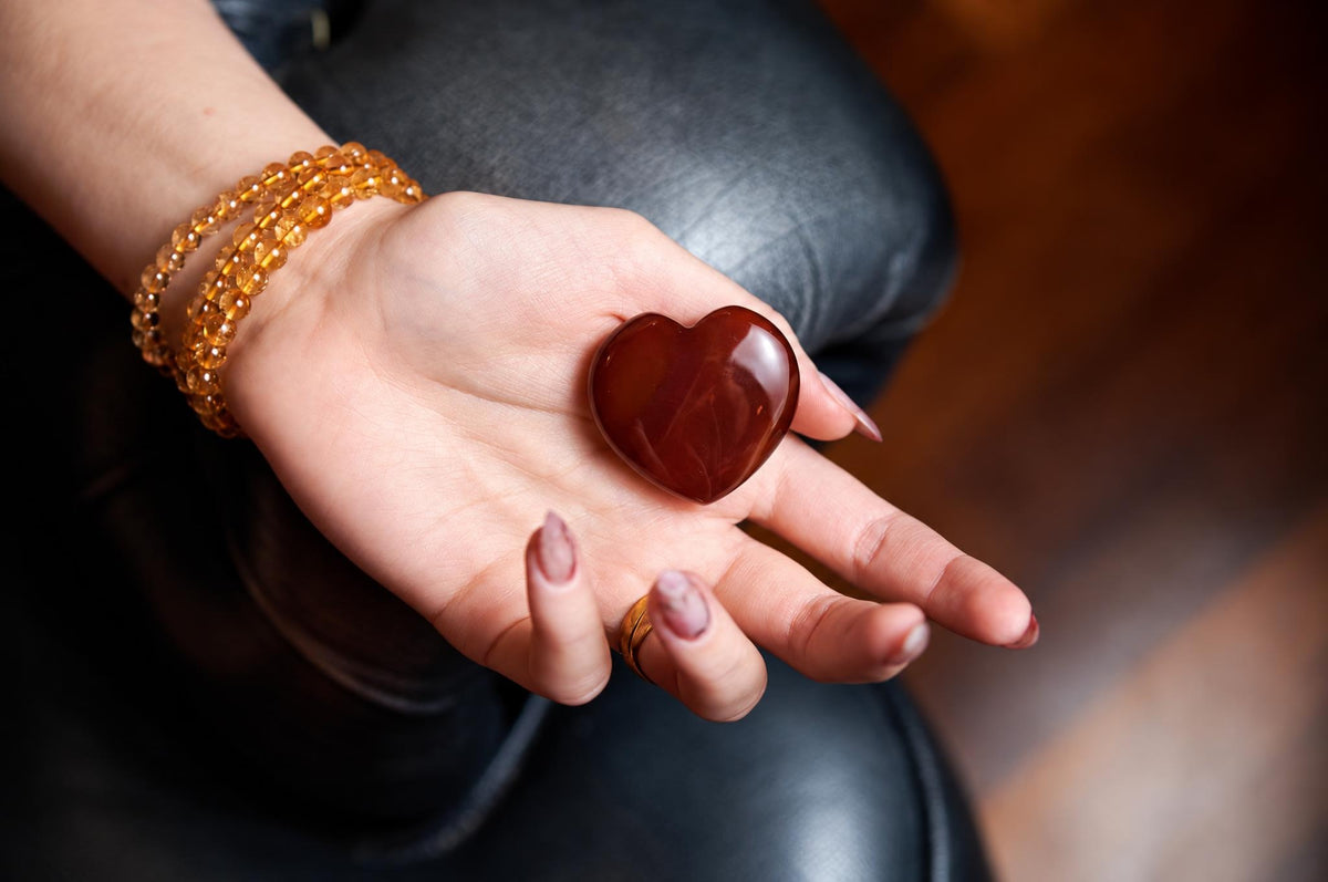 A hand holds a polished, reddish-brown heart-shaped stone on a dark surface.