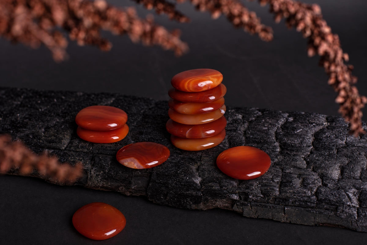 Polished, oval carnelian palmstones in warm orange and red tones are stacked and scattered on a dark wooden surface.