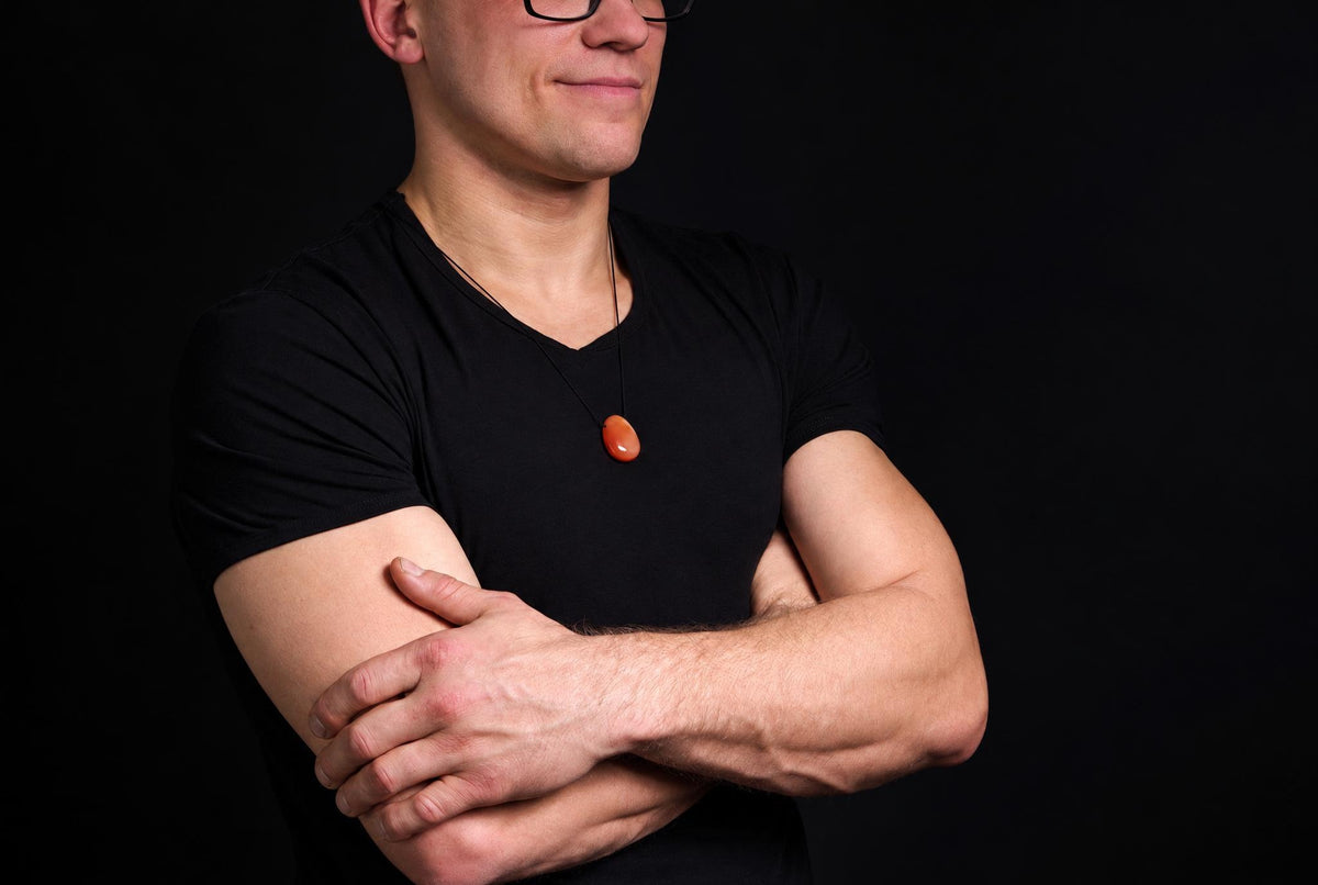 A man in a black shirt wears a necklace with an oval, orange-brown pendant against a dark background.