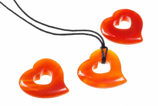 Three heart-shaped pendants in orange and red tones with a central heart cutout, attached to black cords on a white surface.