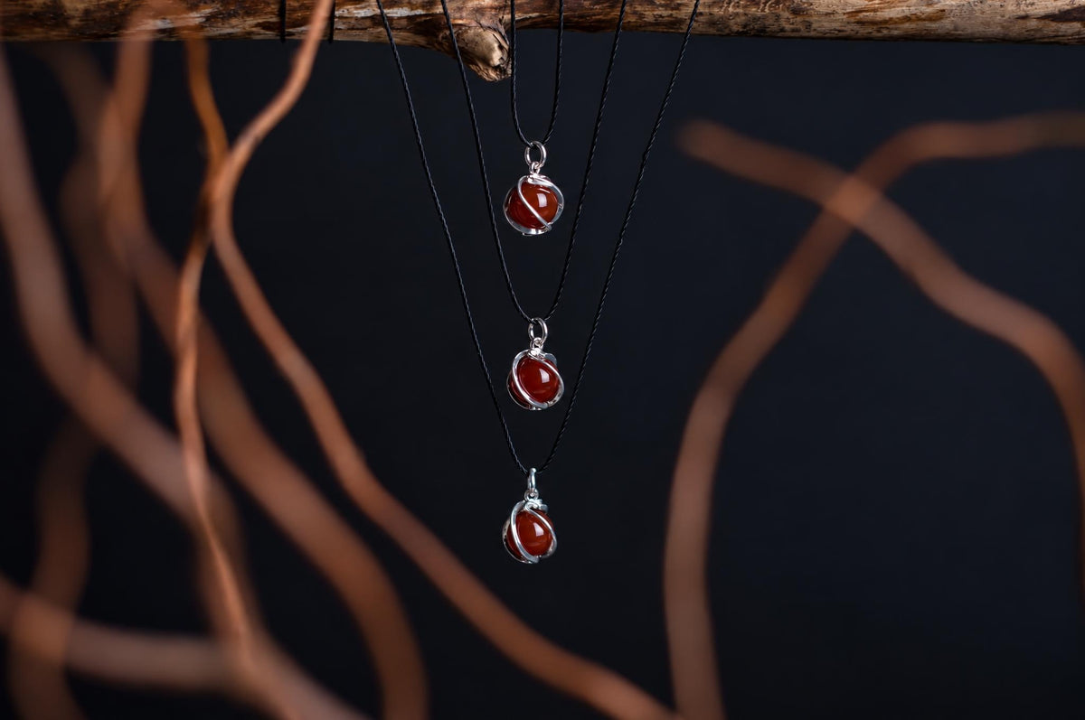 Three silver-toned pendants with red spheres hang from black cords on a dark surface, framed by blurred brown branches.