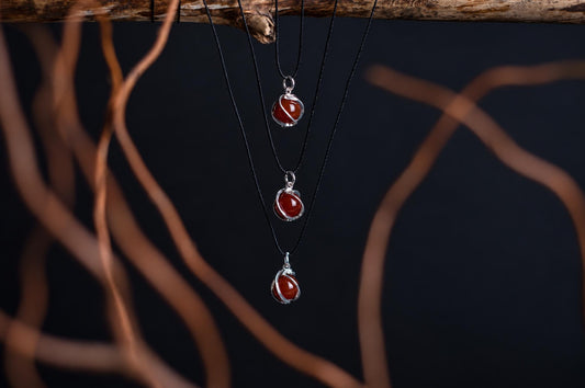 Three silver-toned pendants with red spheres hang from black cords on a dark surface, framed by blurred brown branches.