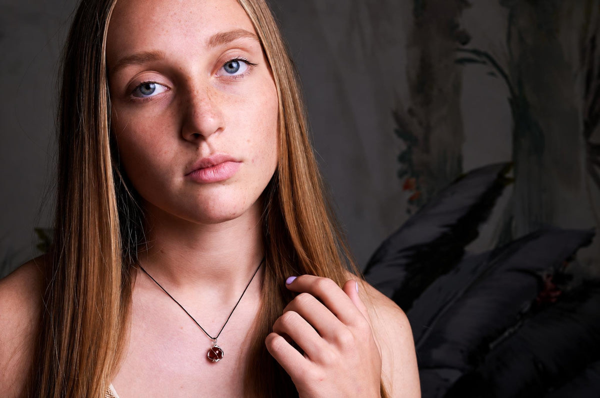 A young woman with long brown hair wears a necklace with a dark red, round pendant on a black cord against a dark background.
