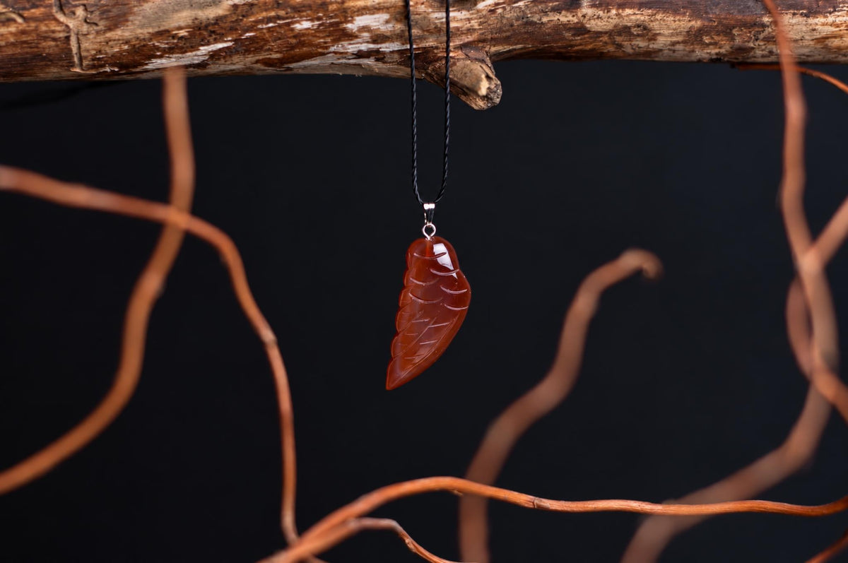 A polished, reddish-brown leaf-shaped pendant with carved veins hangs from a black cord on a dark surface.