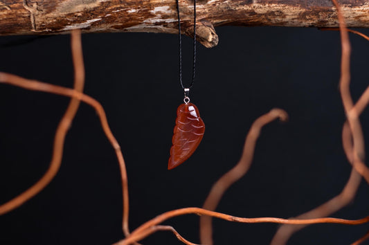 A polished, reddish-brown leaf-shaped pendant with carved veins hangs from a black cord on a dark surface.