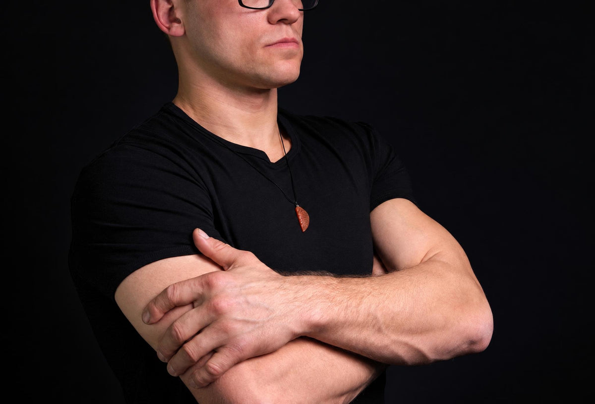 A man in a black t-shirt wears a dark brown, wing-shaped pendant on a thin cord against a dark background.