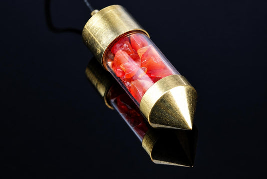 A pendulum with a clear tube filled with red carnelian stones, capped with gold-toned metal ends on a dark surface.