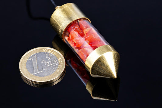A brass pendulum with a clear tube containing red carnelian stones, next to a one-euro coin on a dark reflective surface.