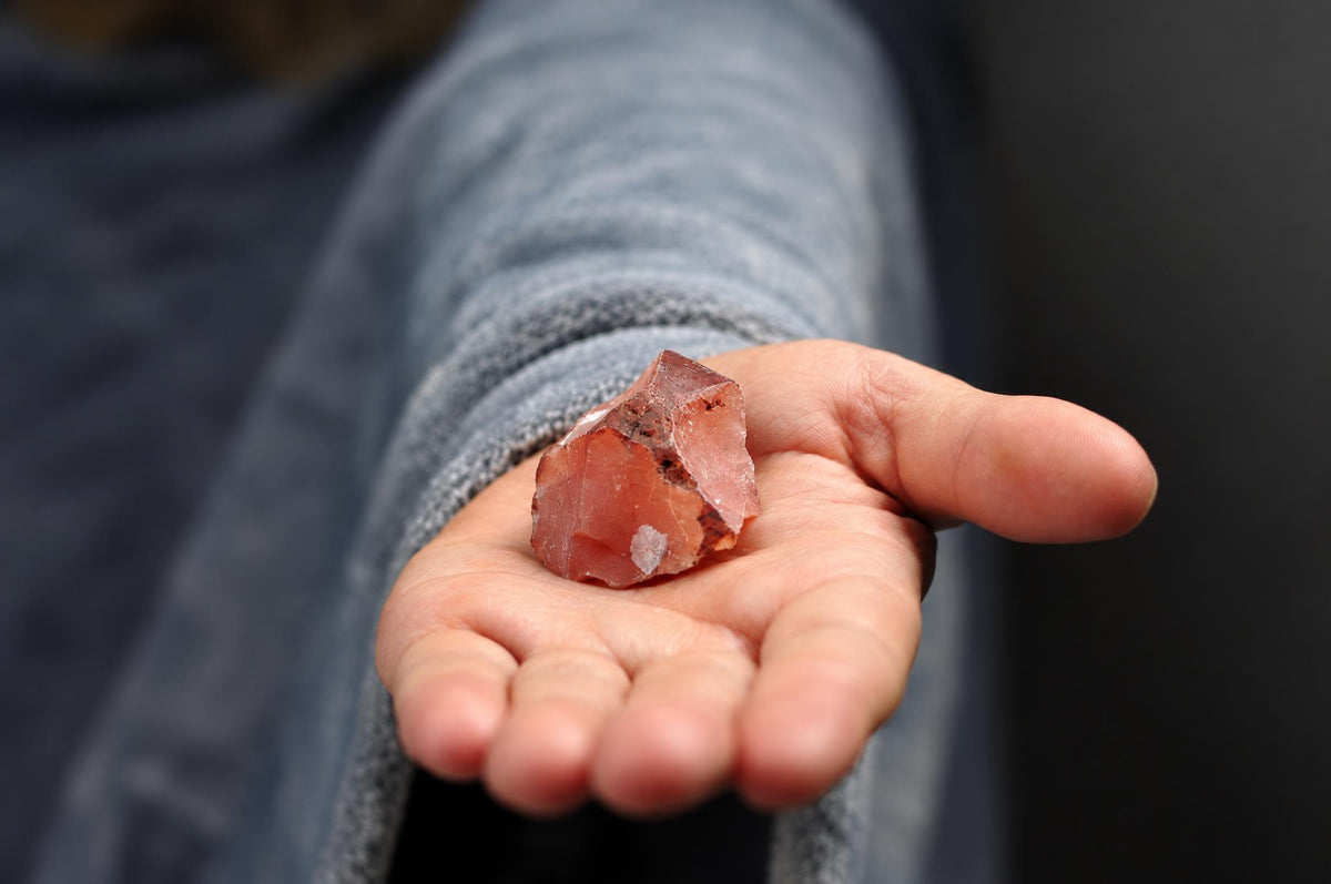 A hand holds a raw, reddish-brown crystal with dark inclusions against a dark surface.