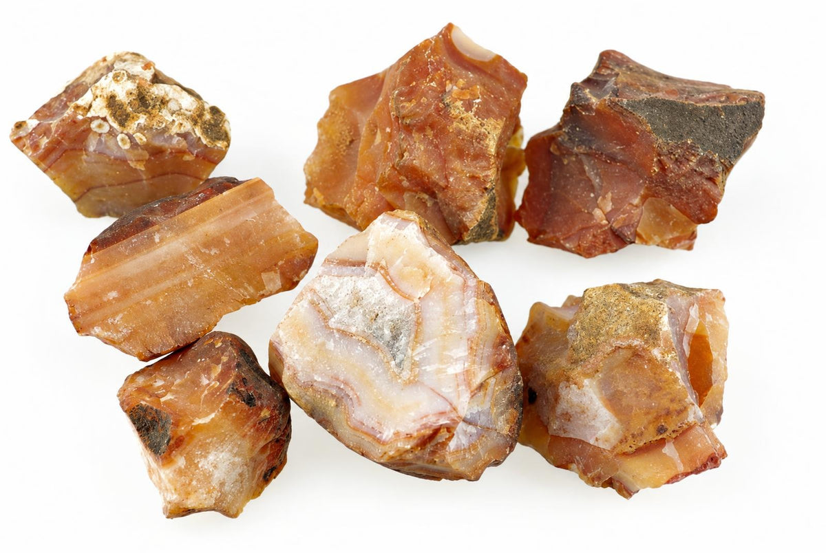 Seven raw carnelian stones in shades of orange, brown and white with dark patches on a pale surface.
