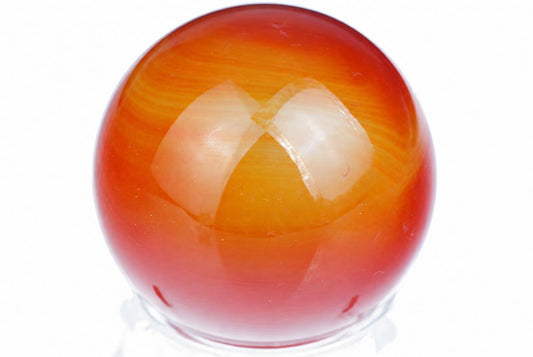 A polished, round carnelian sphere with smooth red and orange bands sits on a clear stand against a white background.