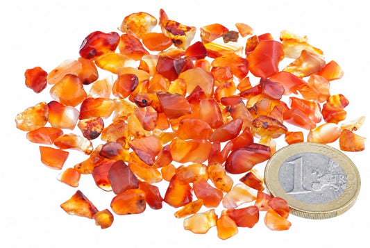 A pile of small, irregular orange and red carnelian stones on a white surface next to a one euro coin.