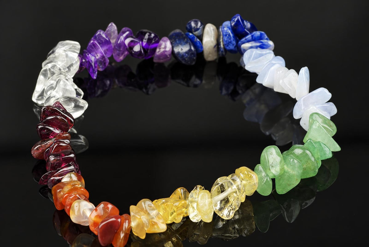 A bracelet made of irregularly shaped chips in clear, purple, blue, white, green, yellow, orange and red stones on a dark reflective surface.