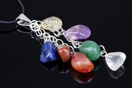 A silver-toned pendant with a black cord holds seven polished, rounded stones in blue, red, orange, green, white, purple and yellow on a dark reflective surface.
