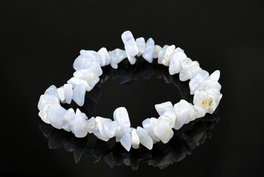 A bracelet made of irregular, polished chalcedony chips in pale blue and white tones rests on a dark reflective surface.