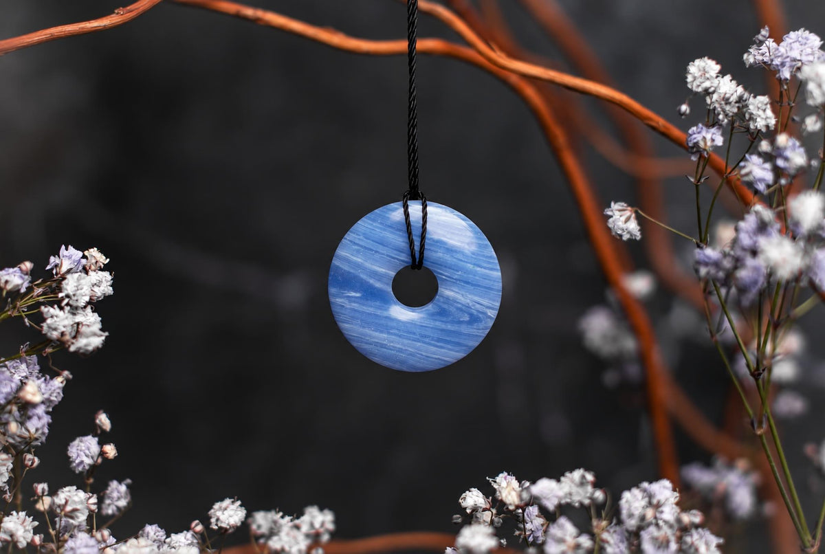 A blue and white swirled donut-shaped pendant hangs from a black cord, surrounded by dried flowers and branches.