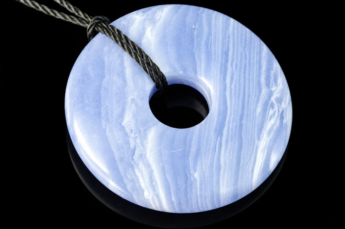 A round, light blue pendant with white swirls and a central hole, threaded on a dark cord against a black background.