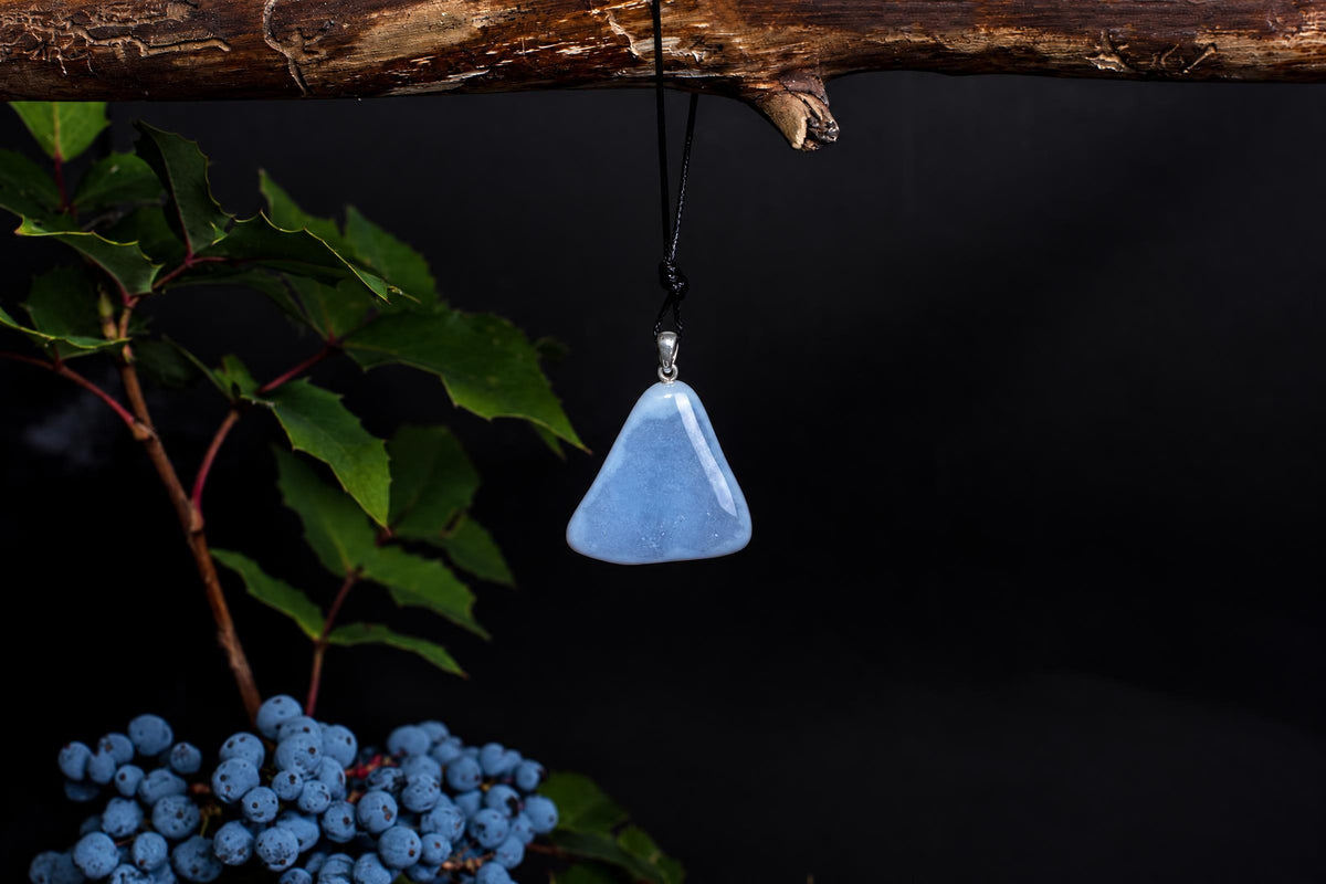 A light blue triangular pendant hangs from a black cord on a dark surface, with green leaves and berries nearby.