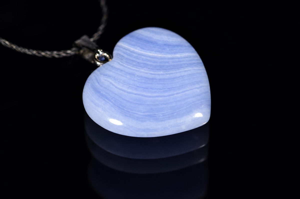 A polished heart-shaped pendant with blue and white banding, attached to a dark cord against a black reflective surface.