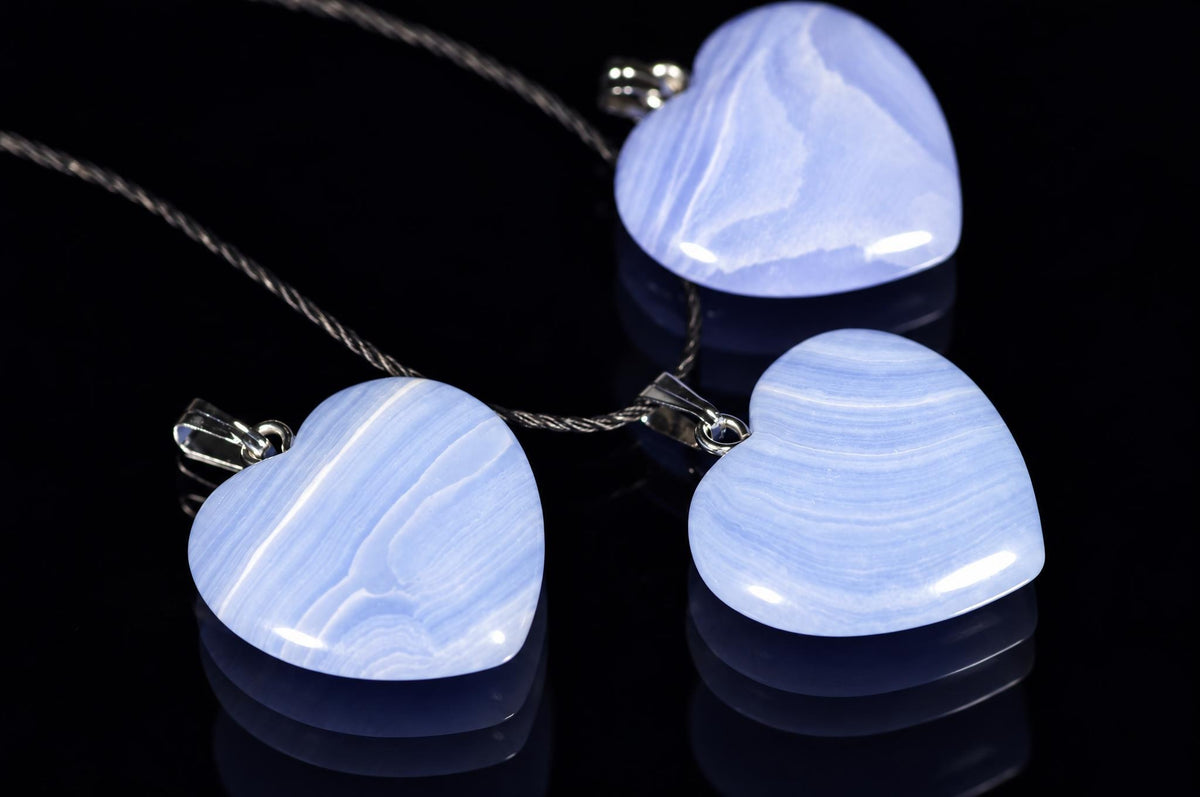 Pale-blue heart-shaped chalcedony pendants with curved white bands lie on black cords against a dark reflective surface.