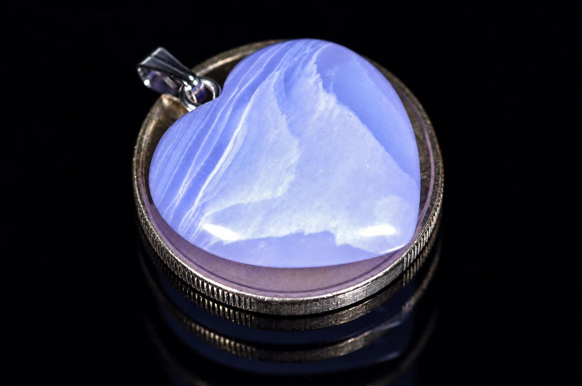 A heart-shaped blue chalcedony pendant with white swirls sits in a silver-toned bezel on a dark reflective surface.