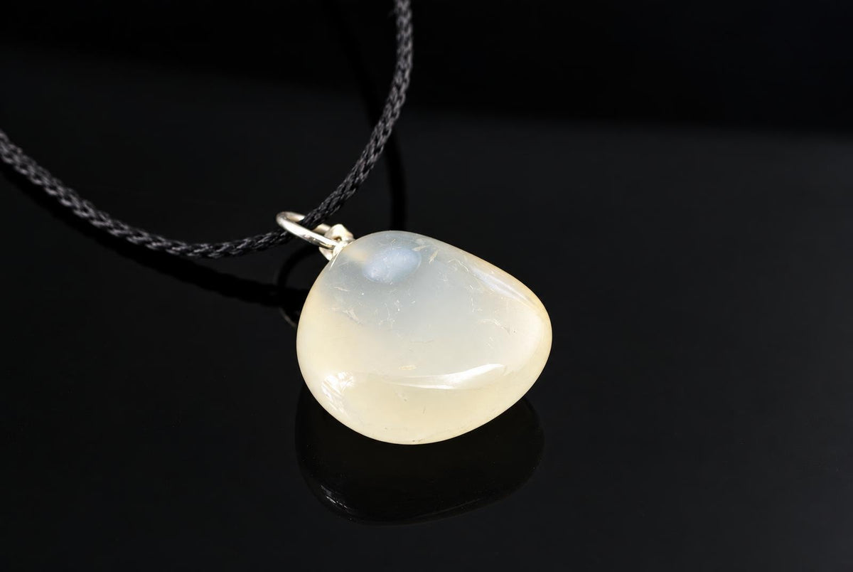 A smooth, pale chalcedony pendant with a silver-toned bail hangs from a dark cord on a black surface.