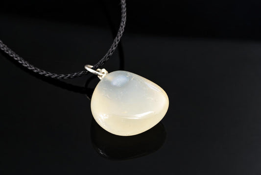 A smooth, pale chalcedony pendant with a silver-toned bail hangs from a dark cord on a black surface.