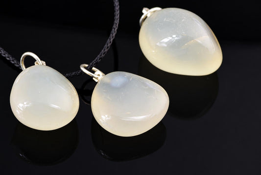 Translucent chalcedony pebble pendants show pale cream and grey colouring, displayed with metallic loops and a black cord on a dark surface.