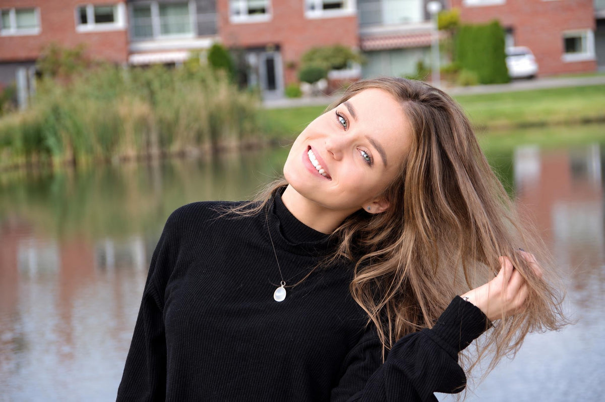A smiling woman with long brown hair wears a black turtleneck and a silver-toned pendant necklace, holding her hair near a lake.