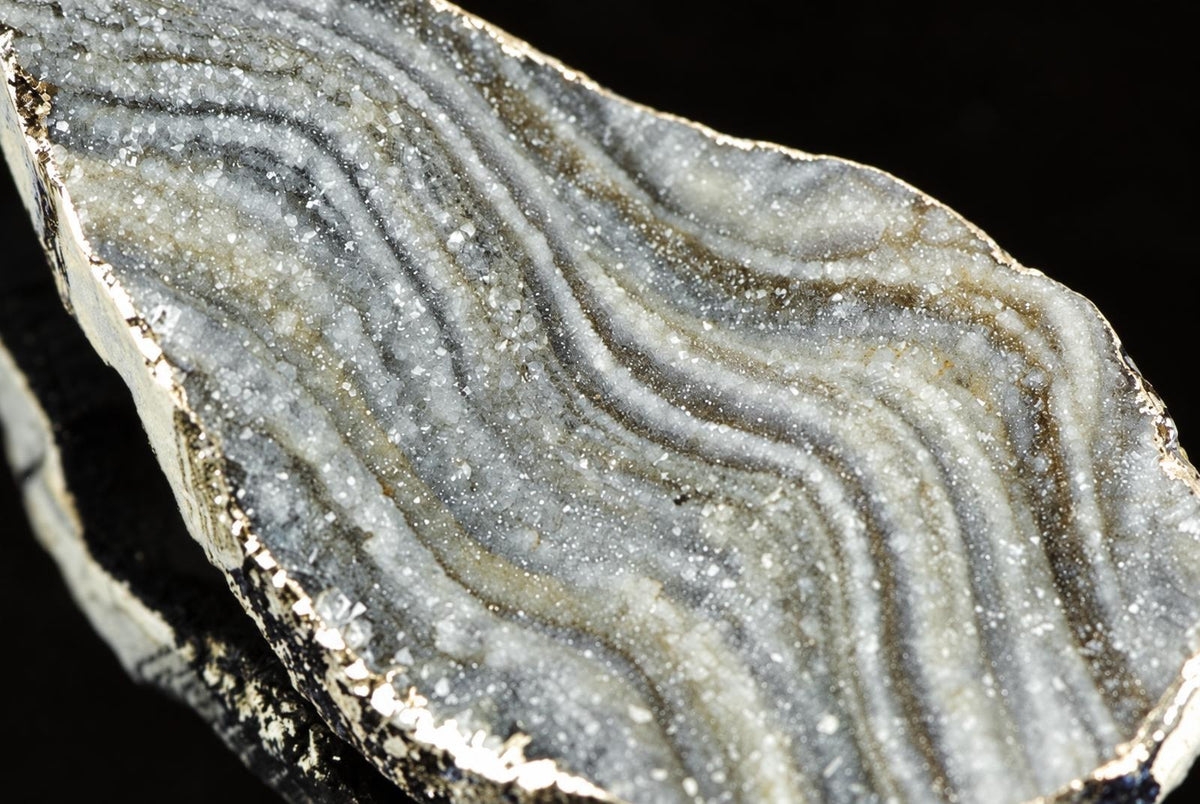 A polished chalcedony slice with wavy bands of white, grey and brown crystals on a dark surface.