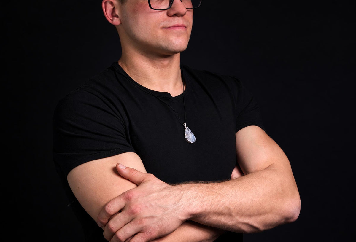 A man in a black t-shirt wears a silver-toned pendant with an irregular, pale blue-grey stone on a dark surface.