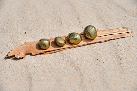 Four smooth, metallic golden-green stones sit in a row on a strip of wood over pale sand.