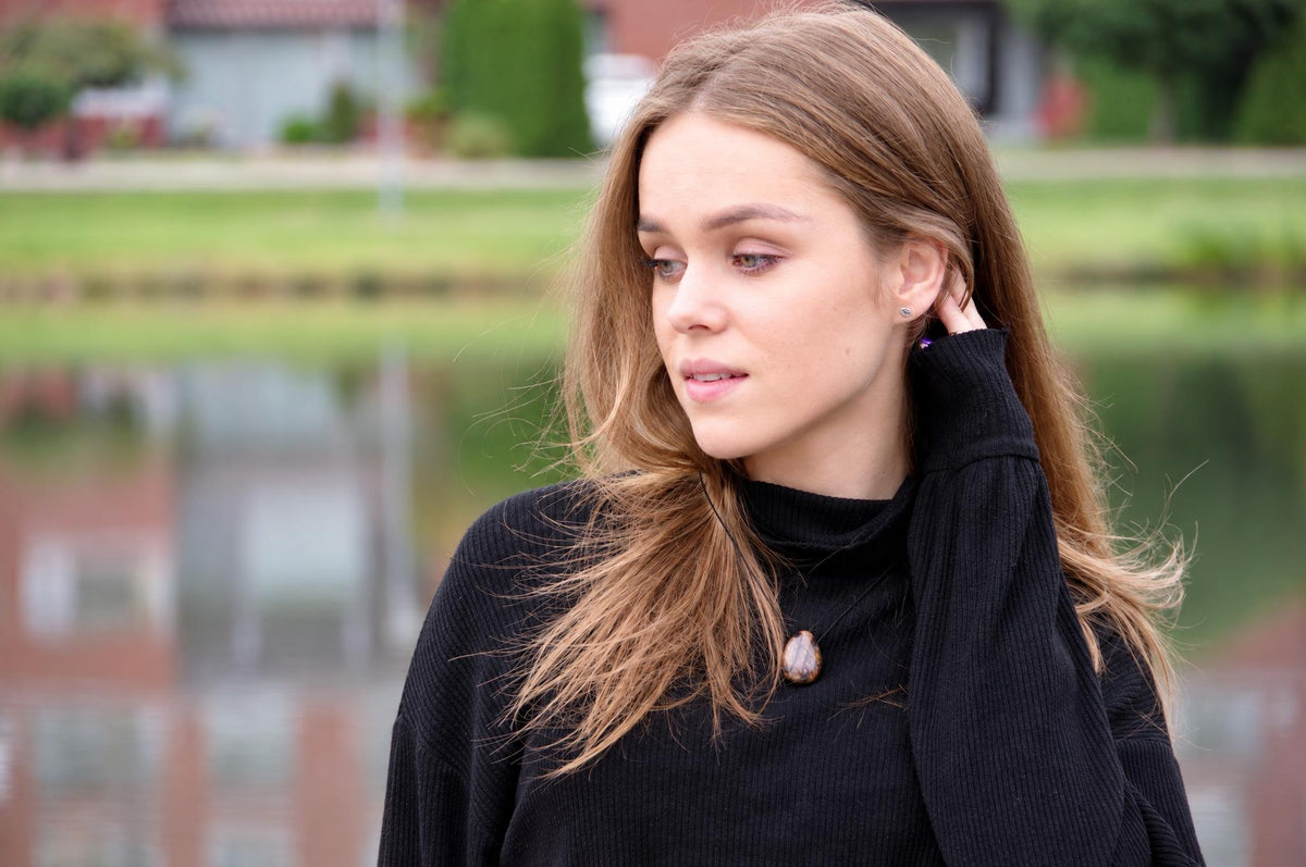 A woman wears a black turtleneck sweater with a brown, teardrop-shaped pendant necklace against a blurred outdoor background.