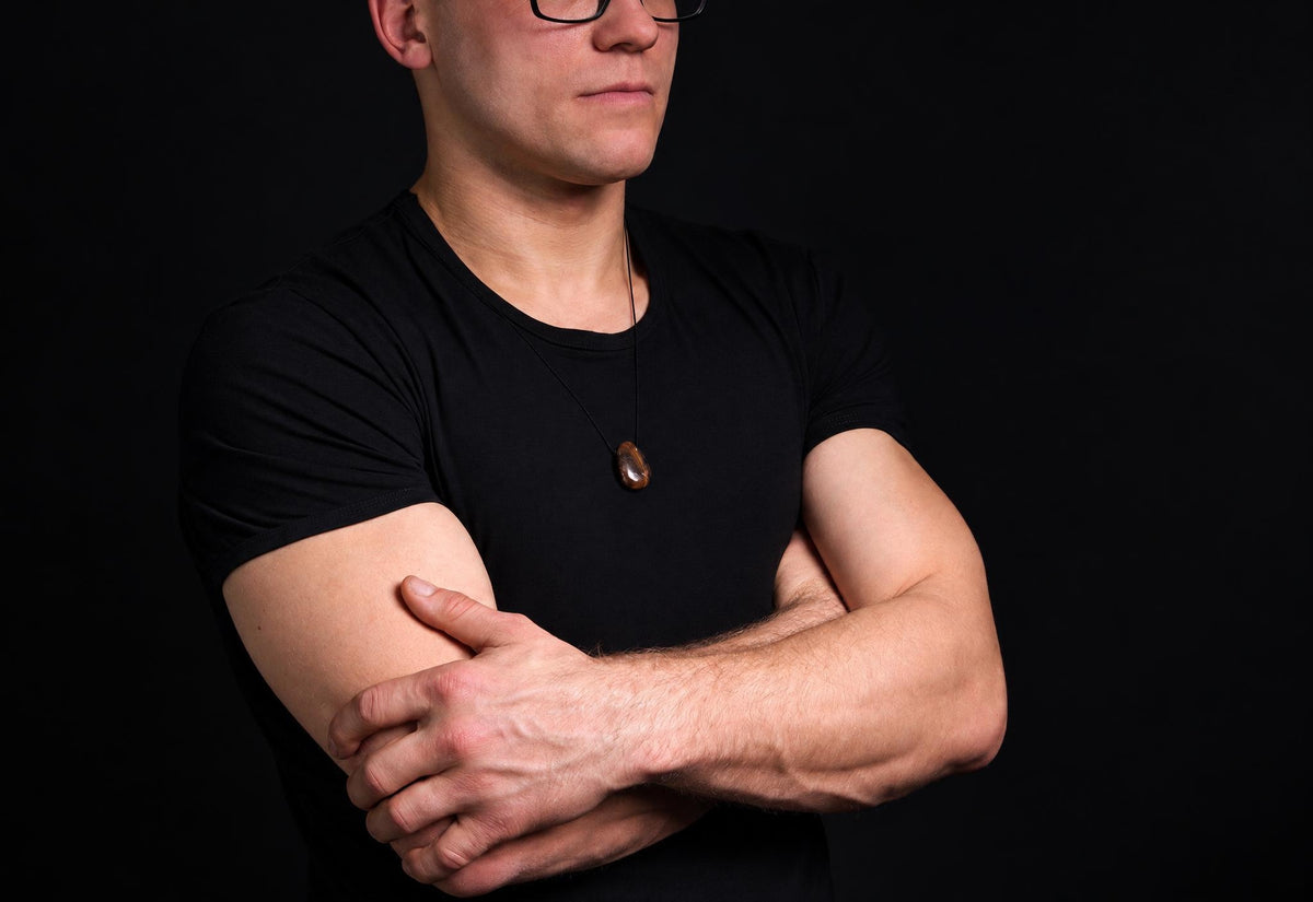 A man in a black t-shirt wears a pendant with a dark brown, polished drop-shaped stone on a thin cord against a dark background.