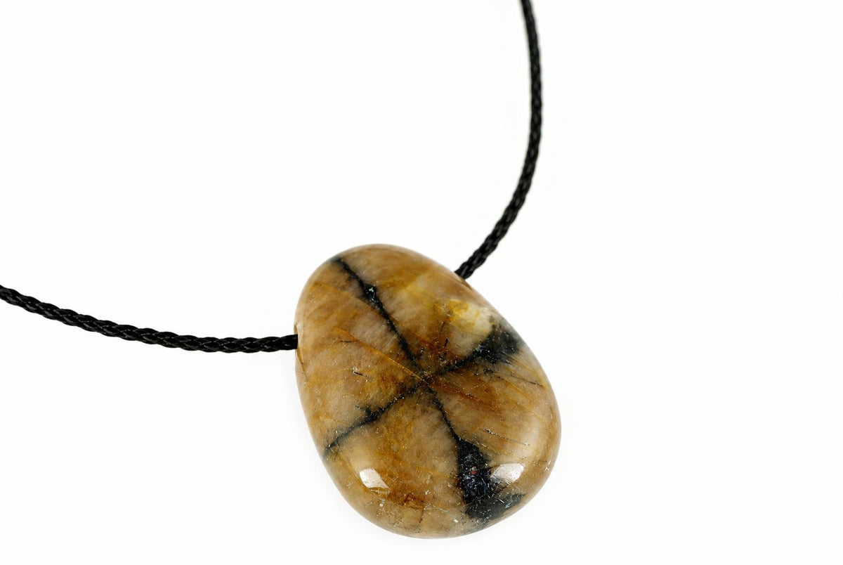 A polished Chiastolite pendant with a smooth, oval shape and dark brown to black inclusions on a pale surface, hanging from a black cord against a white background.
