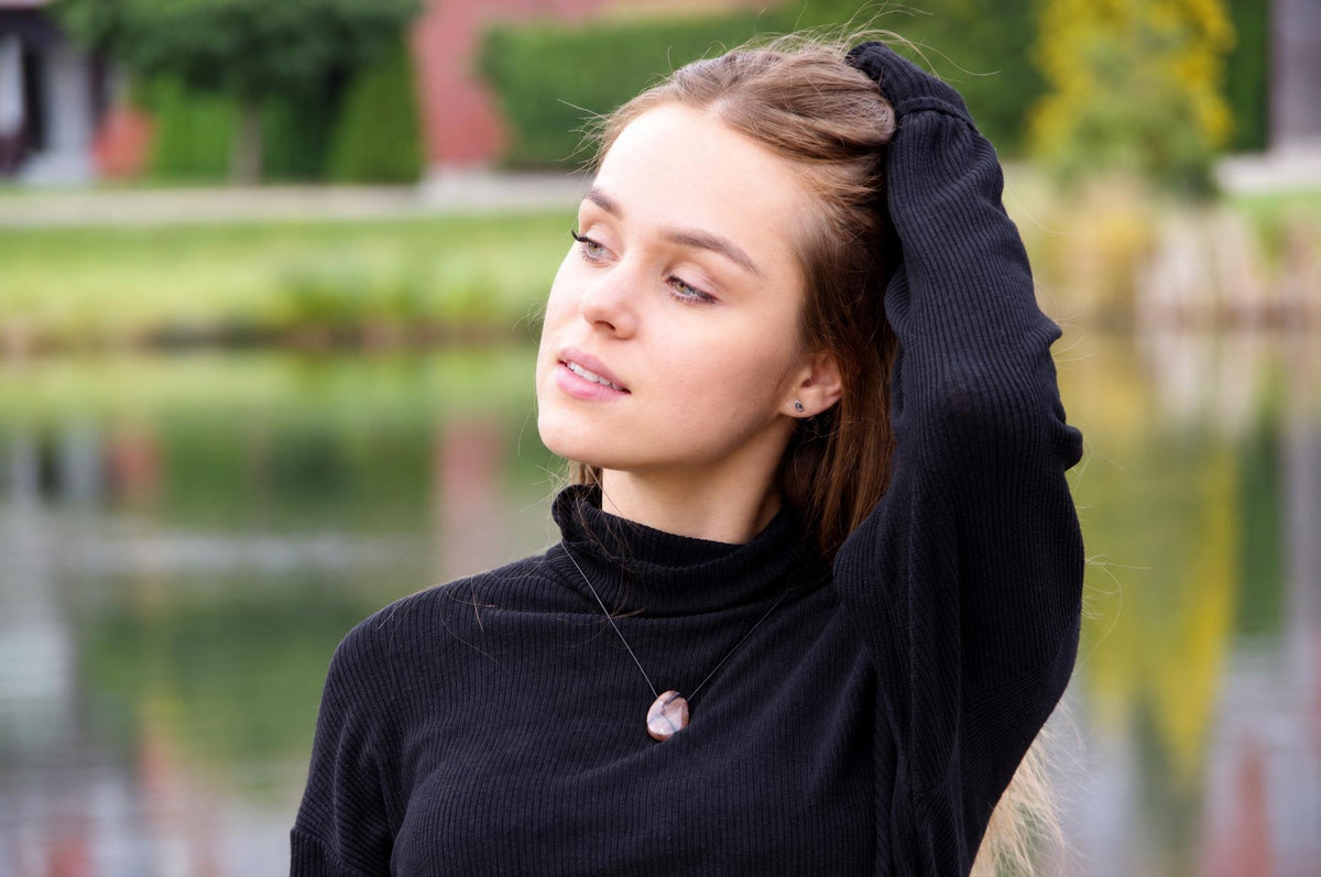 A woman wears a black turtleneck with a round, pinkish pendant on a thin cord against a blurred outdoor background.