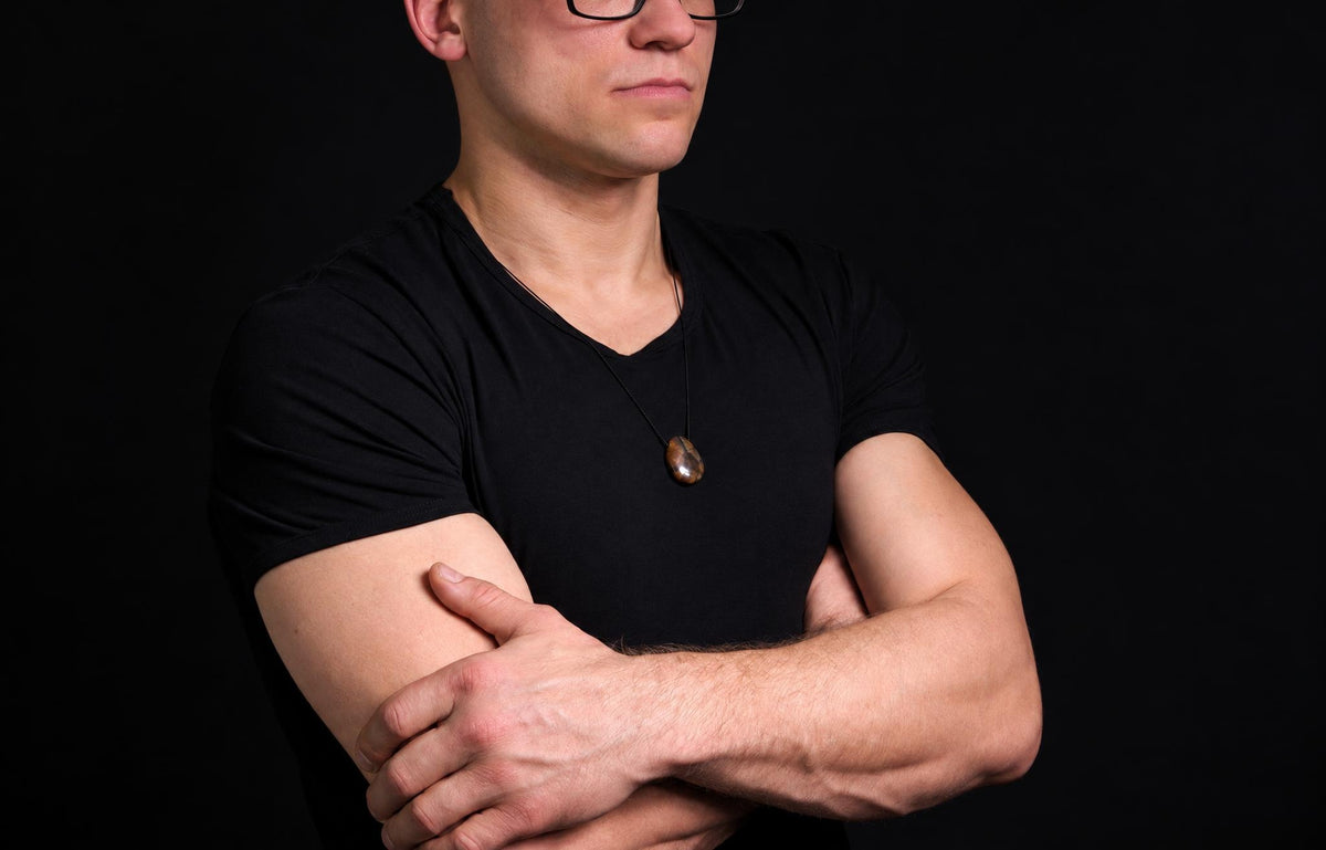 A man in a black t-shirt wears a pendant with a dark brown, oval stone on a thin cord against a dark background.