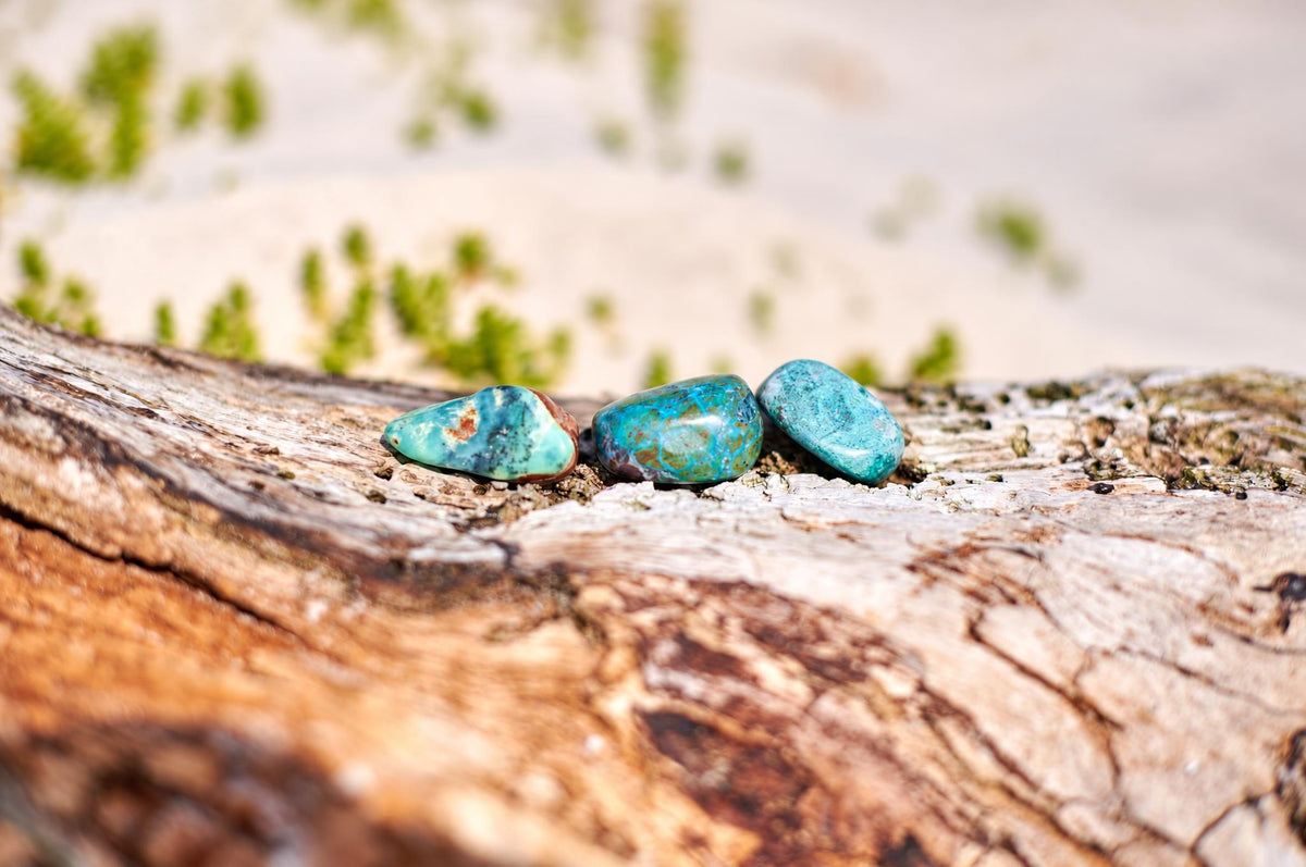 Small polished chrysocolla stones with blue green colouring and brown patches rest on a weathered wooden surface in an outdoor setting.