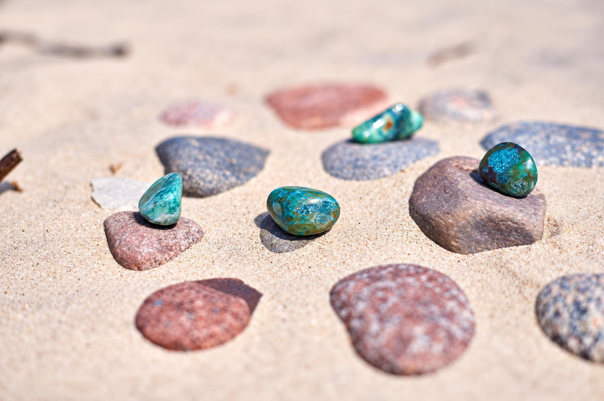 Small blue and green chrysocolla stones rest among reddish and gray pebbles on sand, with rounded surfaces and mottled darker patches.